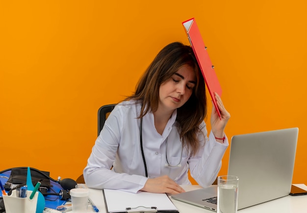 닫힌 된 눈으로 청진 기 의료 가운을 입고 피곤 된 중년 여성 의사는 오렌지 벽에 머리에 폴더를 넣어 의료 도구와 노트북에 노트북에서 작동
