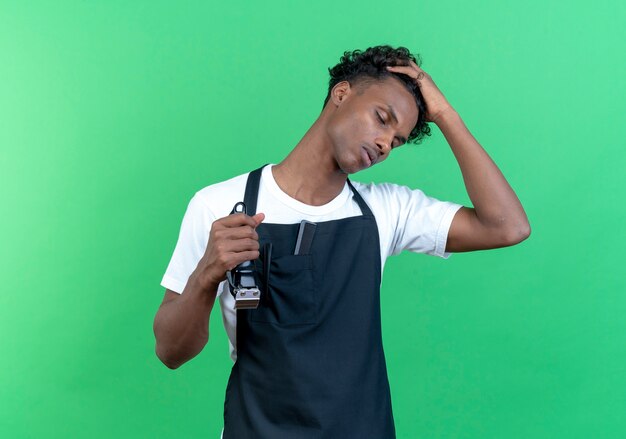 With closed eyes tired young afro-american male barber wearing uniform putting hand on head and holding hair clippers isolated on green wall