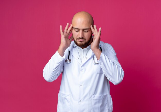 With closed eyes tired young bald male doctor wearing medical robe and stethoscope putting fingers on forehead isolated on pink with copy space