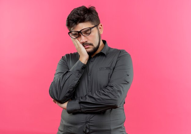 With closed eyes tired young businessman wearing glasses keeping hand on cheek isolated on pink