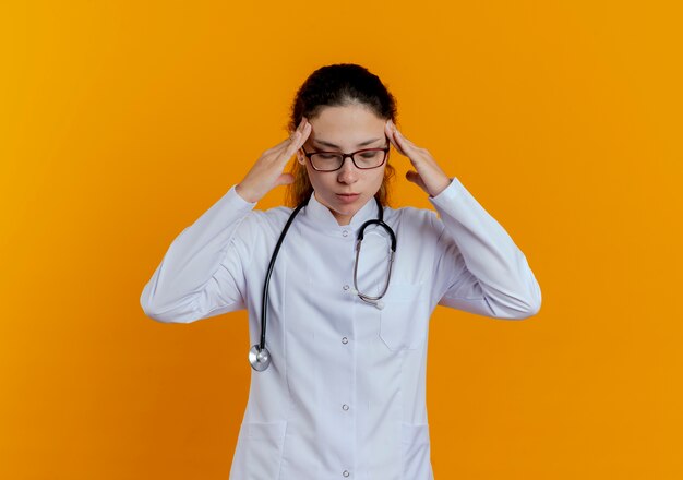 目を閉じて疲れた若い女性医師が医療ローブと眼鏡をかけてこめかみに手を置いて聴診器を分離