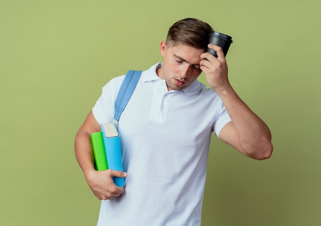 With closed eyes tired young handsome male student wearing back bag holding books and putting cup of coffee on forehead isolated on olive green