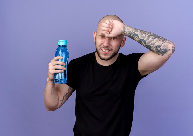 С закрытыми глазами усталый молодой спортивный мужчина держит бутылку с водой и кладет запястье на лоб, изолированный на фиолетовом