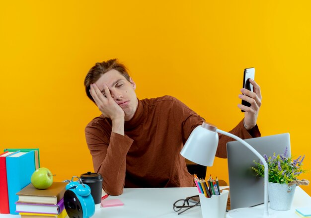 With closed eyes tired young student boy sitting at desk with school tools holding phone and covered face on yellow