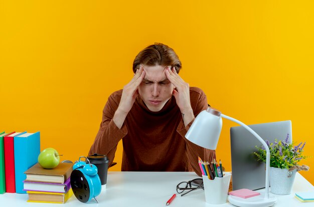 With closed eyes tired young student boy sitting at desk with school tools putting hand around eyes isolated on yellow wall