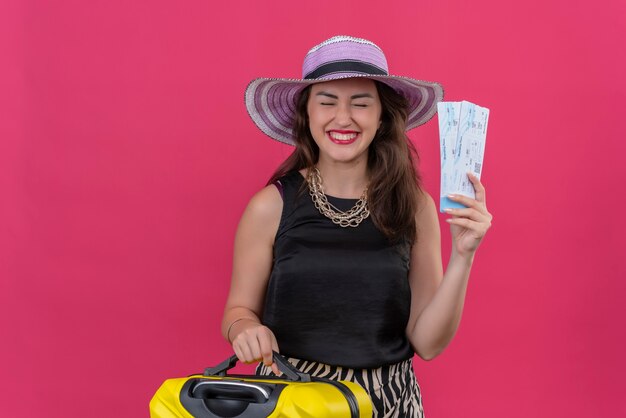 With closed eyes traveler young girl wearing black undershirt in hat holding tickets on red background