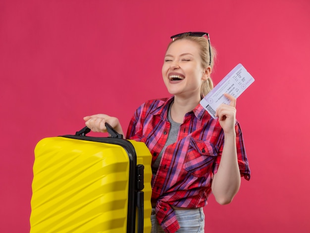 With closed eyes traveler young girl wearing red shirt in glasses holding suitcase and ticket on isolated pink background