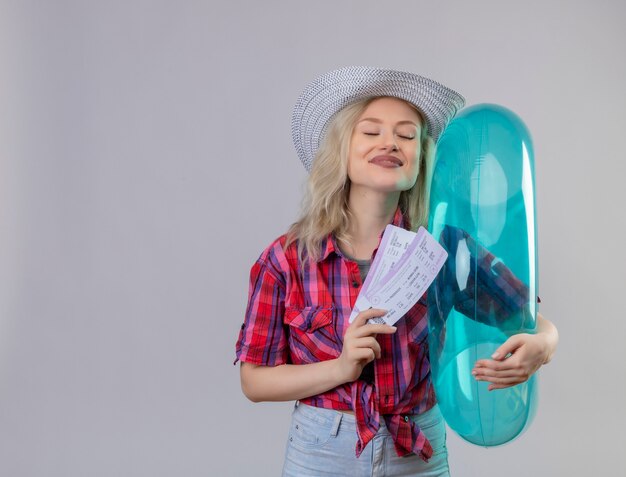 With closed eyes traveler young girl wearing red shirt in hat holding inflatable ring and tickets on isolated white background