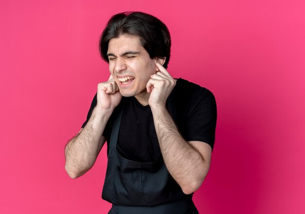 With closed eyes unpleased young handsome male barber in uniform closing ears with fingers isolated on pink