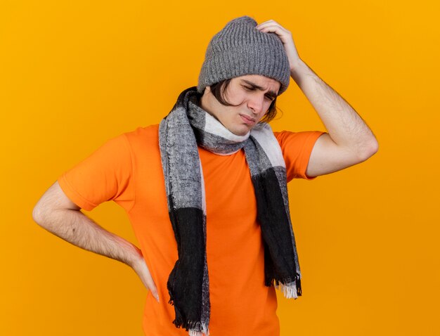 С закрытыми глазами недовольный молодой больной в зимней шапке с шарфом кладет руки на больную голову и бедро, изолированные на оранжевом фоне