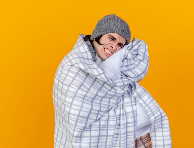 With closed eyes unpleased young ill man wearing winter hat with scarf wrapped in plaid isolated on orange