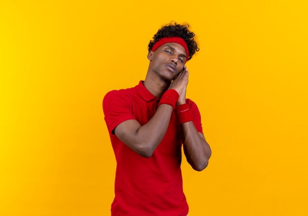 With closed eyes young afro-american sporty man wearing headband and wristband showing sleep gesture isolated on yellow wall