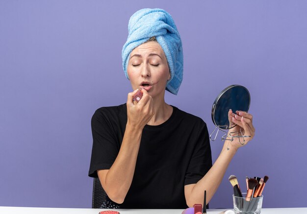 With closed eyes young beautiful girl sits at table with makeup tools wiping hair in towel applying lipstick holding mirror isolated on blue background