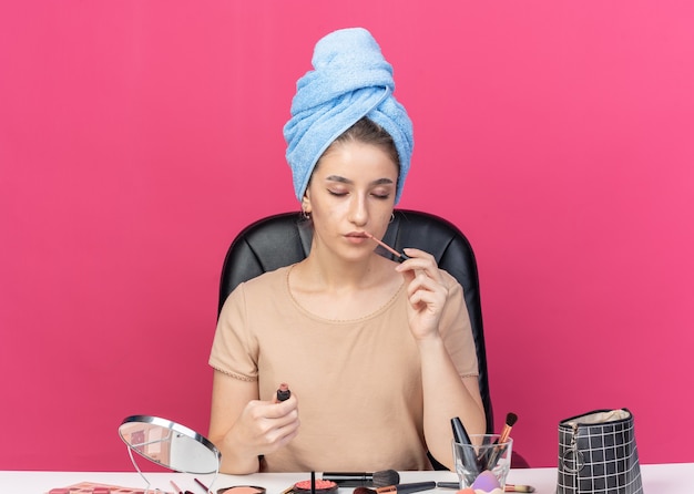 With closed eyes young beautiful girl sits at table with makeup tools wrapped hair in towel applying lipgloss isolated on pink background Free Photo