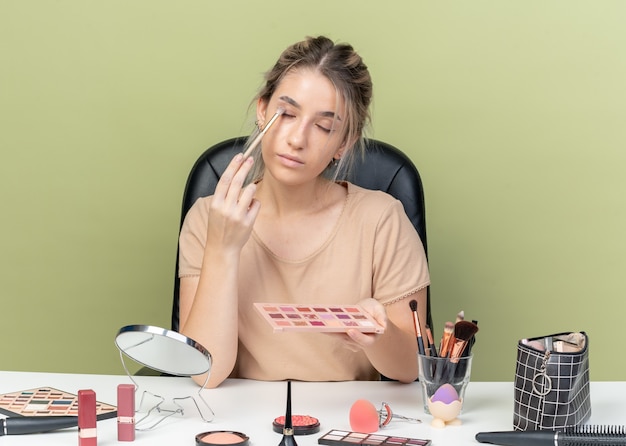 目を閉じて、オリーブグリーンの背景に分離された化粧ブラシでアイシャドウを適用する化粧ツールで机に座っている若い美しい少女