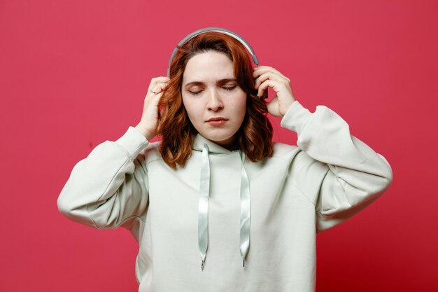 With closed eyes young beautiful girl in white sweater wearing headphones isolated on pink background