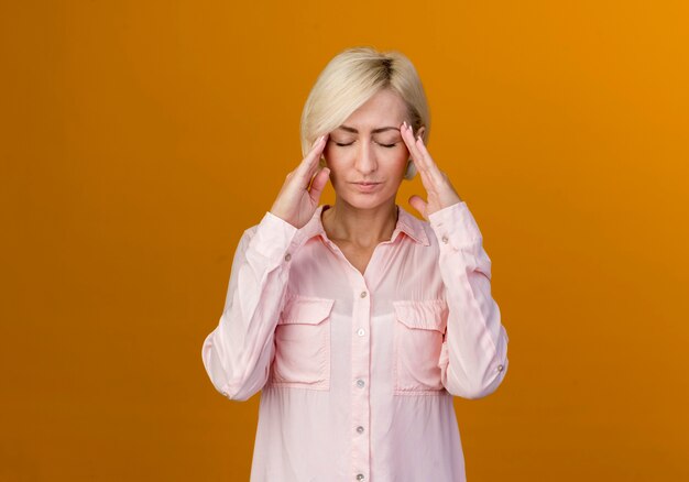 With closed eyes young blonde slavic woman putting hands on temple isolated on orange wall