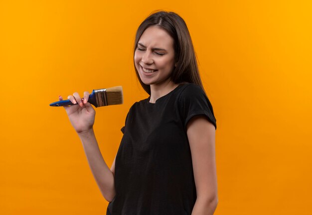 With closed eyes young caucasian girl wearing black t-shirt holding paint brush and pretend sing on isolated orange wall