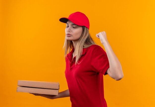 Молодая доставщица с закрытыми глазами в красной форме и кепке держит коробку из-под пиццы и показывает жест "да", изолированный на оранжевой стене