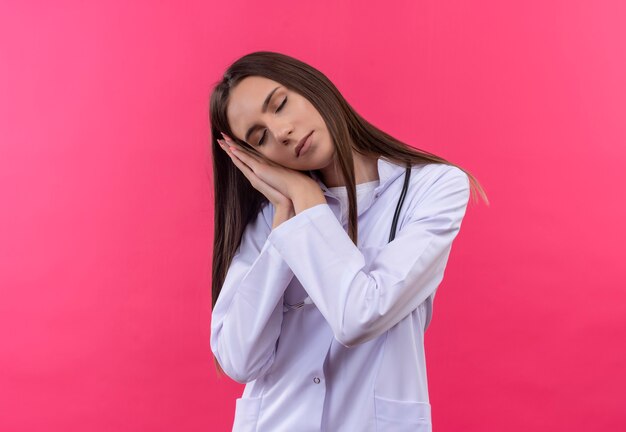 Молодая девушка-врач с закрытыми глазами в медицинском халате со стетоскопом показывает жест сна на изолированной розовой стене