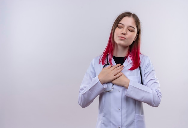 Foto gratuita con gli occhi chiusi giovane ragazza medico che indossa stetoscopio abito medico mettendo la mano sul petto sulla parete bianca isolata con lo spazio della copia