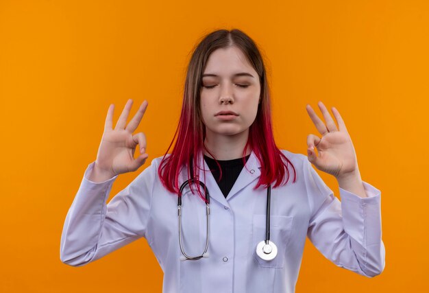 With closed eyes young doctor girl wearing stethoscope medical robe showing okey gesture on isolated orange wall