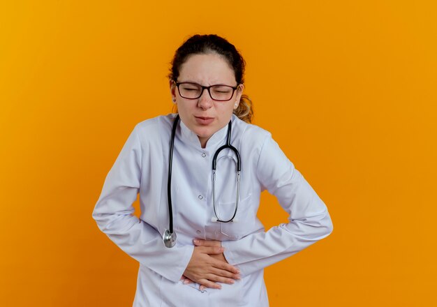 With closed eyes young female doctor wearing medical robe and stethoscope with glasses putting hands on aching stomach isolated