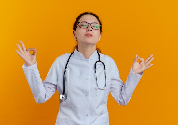 Foto gratuita con gli occhi chiusi giovane medico femminile che indossa veste medica e stetoscopio con gli occhiali che mostrano la meditazione isolata sulla parete arancione