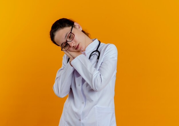 With closed eyes young female doctor wearing medical robe and stethoscope with glasses showing sleep gesture isolated