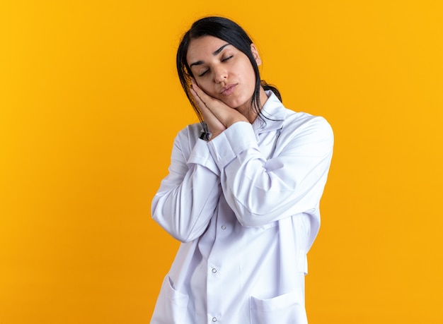 With closed eyes young female doctor wearing medical robe with stethoscope showing sleep gesture isolated on yellow background