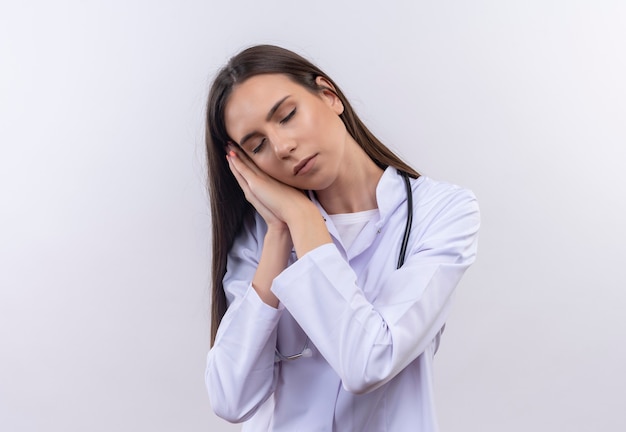 Молодая девушка с закрытыми глазами в медицинском халате со стетоскопом показывает жест сна на изолированном белом