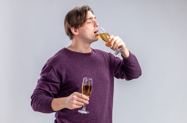 With closed eyes young guy on valentines day holding and drinks glasses of champagne isolated on white background