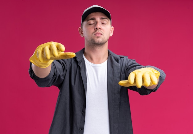 With closed eyes young hamdsome cleaning guy wearing t-shirt and cap with gloves holding out hands  isolated on pink wall