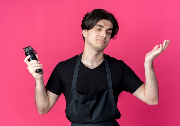 With closed eyes young handsome male barber in uniform holding hair clippers and spread hand isolated on pink