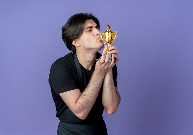 With closed eyes young handsome male barber in uniform holding and kissing winner cup isolated on blue