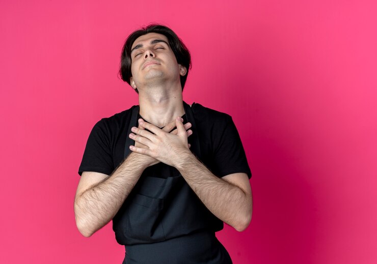 Free Photo with closed eyes young handsome male barber in uniform putting hands on chest isolated on pink