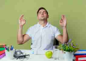 Foto gratuita con gli occhi chiusi giovane allievo maschio bello che si siede allo scrittorio con gli strumenti della scuola che mostrano il gesto di meditazione isolato su verde oliva