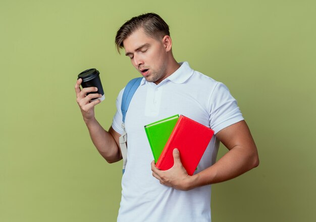 With closed eyes young handsome male student wearing back bag holding cup of coffee and books isolated on olive green