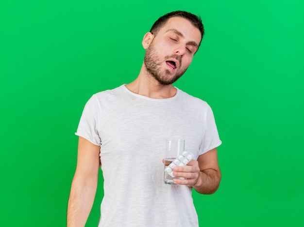 Бесплатное фото С закрытыми глазами молодой больной мужчина держит таблетки со стаканом воды, изолированные на зеленом фоне