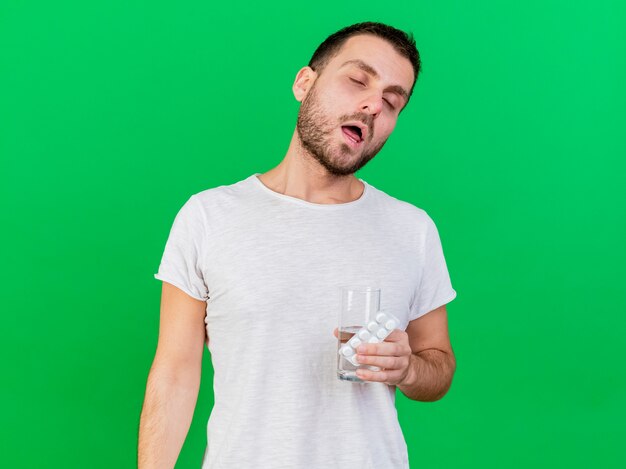 With closed eyes young ill man holding pills with glass of water isolated on green background