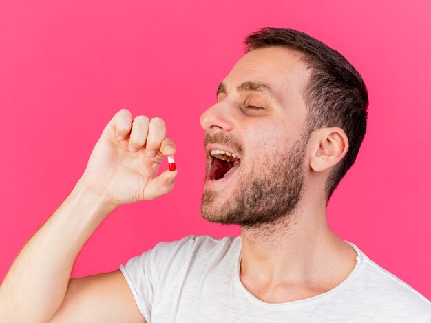 With closed eyes young ill man putting pills in mouth isolated on pink background