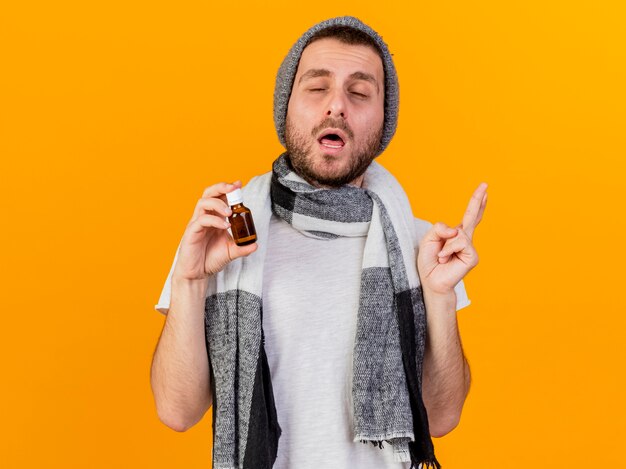 With closed eyes young ill man wearing winter hat and scarf holding medicine in glass bottle isolated on yellow background