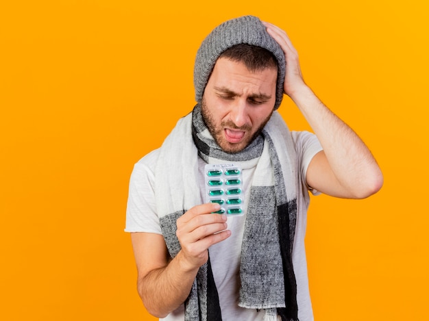 With closed eyes young ill man wearing winter hat and scarf holding pills and showing tongue putting hand on head isolated on yellow background