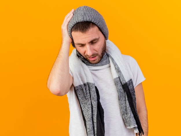 With closed eyes young ill man wearing winter hat and scarf putting hand on aching head isolated on yellow background