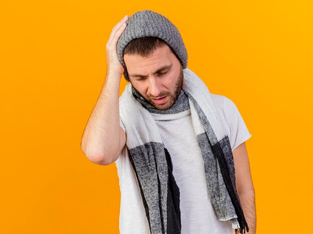 With closed eyes young ill man wearing winter hat and scarf putting hand on aching head isolated on yellow background