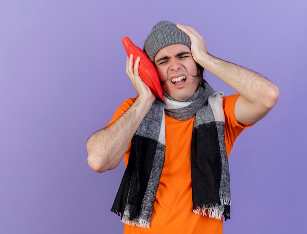 With closed eyes young ill man wearing winter hat with scarf putting hot water bag on cheek putting hand on aching head isolated on purple background