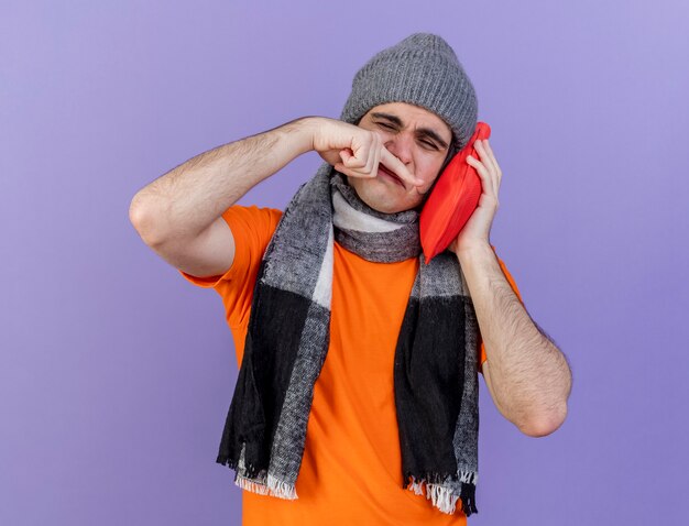 With closed eyes young ill man wearing winter hat with scarf putting hot water bag on cheek wiping nose with hand isolated on purple background