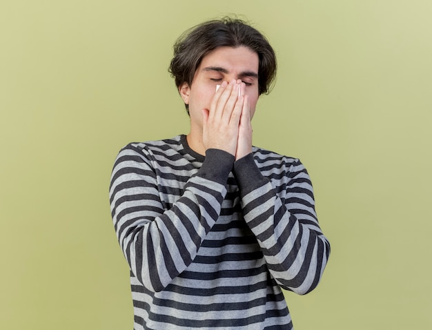 With closed eyes young ill man wiping nose with nose isolated on olive green background