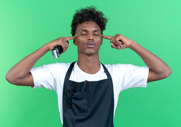 With closed eyes young male barber wearing uniform holding hair clippers