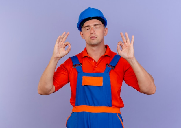 With closed eyes young male builder wearing uniform and safety helmet showing okey gesture on purple